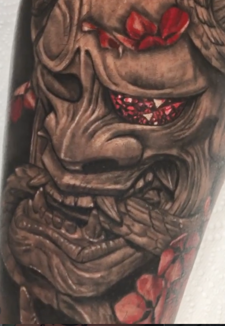 Close detail of a black-and-grey Hannya mask tattoo with red accents