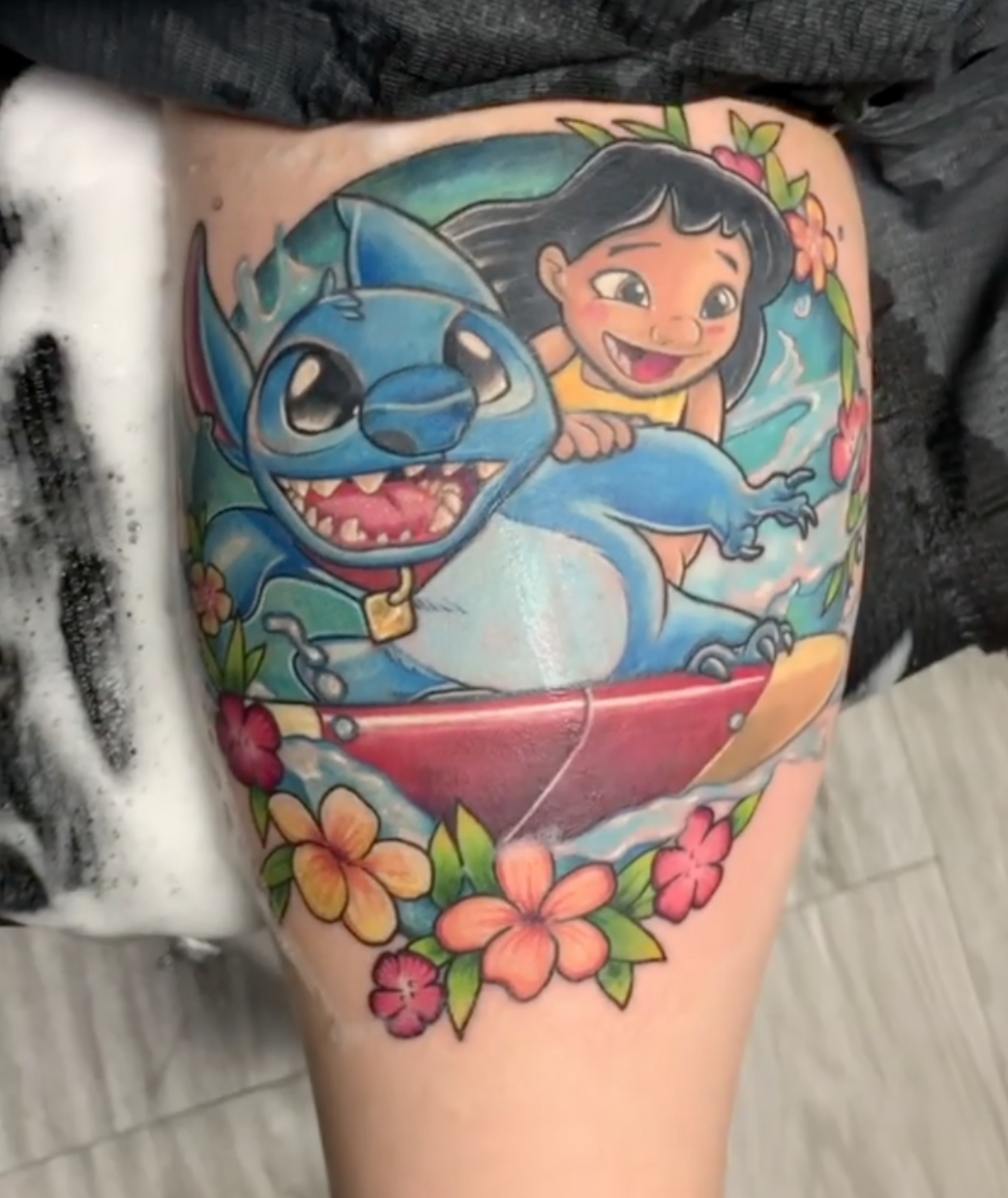 Color Lilo and Stitch surfing tattoo surrounded by tropical flowers